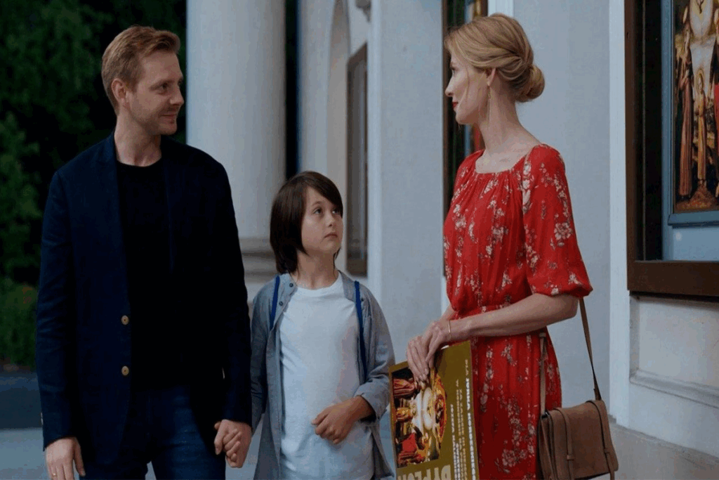 Pais e filho em corredor de galeria de arte, o homem está usando cabelo curto claro, usando camiseta e blazer azul escurto e calça jeans azul, com um menino com cabelo liso, curto, usando camiseta branca e camisa cinza e uma mulher com cabelo loiro em coque  e vestido vermelho estampado