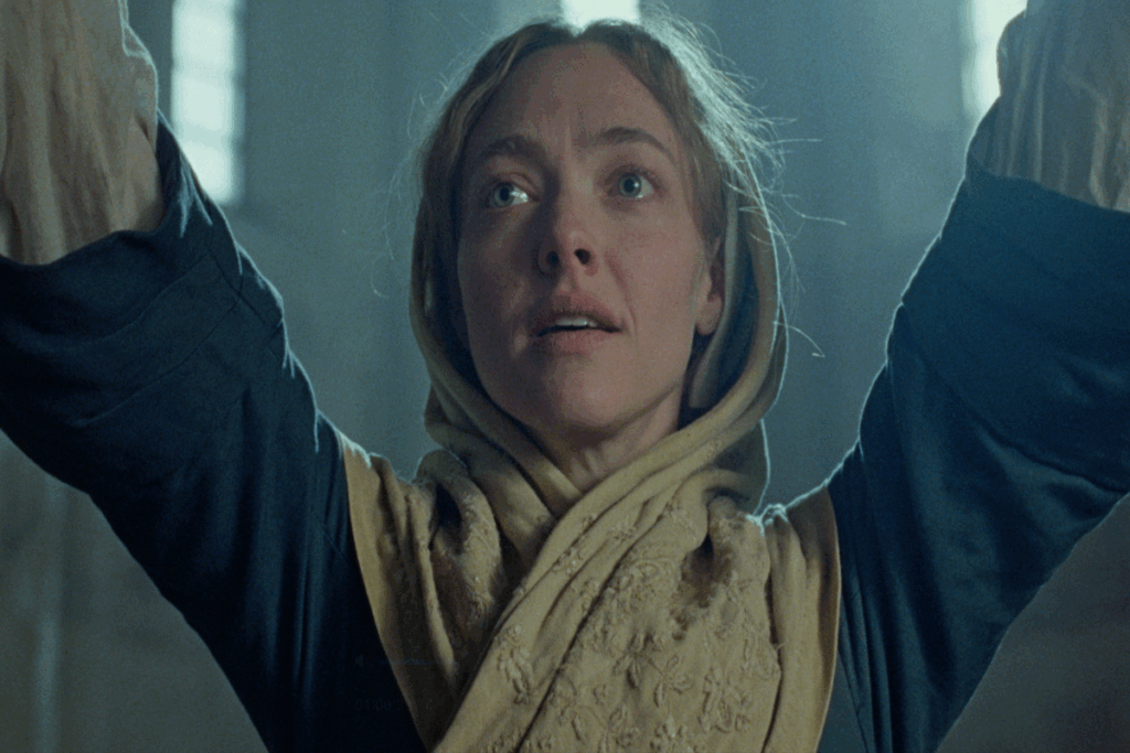 Mulher com cabelo loiro, com lenço bege no cabelo, e camisa de manga longa azul escura com os braços para cima