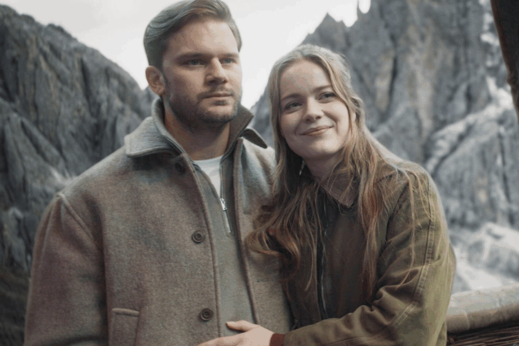 Casal posando para foto, usando casacos em tons de bege e verde, o homem está com um rosto sério e a mulher sorrindo 