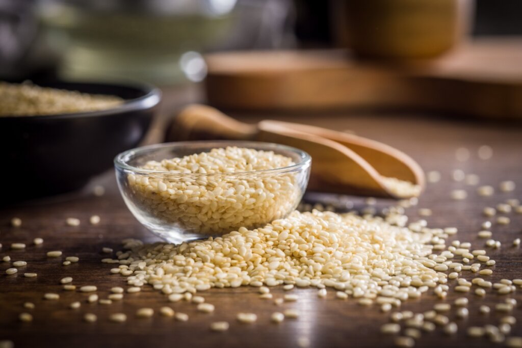 A imagem mostra um pequeno recipiente de vidro cheio de sementes de gergelim sobre uma mesa de madeira. Algumas sementes estão espalhadas ao redor, e ao fundo aparece uma colher de madeira e outros recipientes com o mesmo ingrediente.