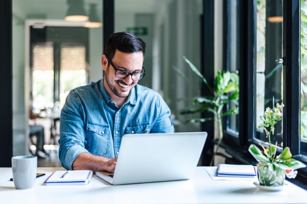 Homem com cabelo curto, barba, usando óculos preto e camisa jeans, mexendo em notebook em escritório