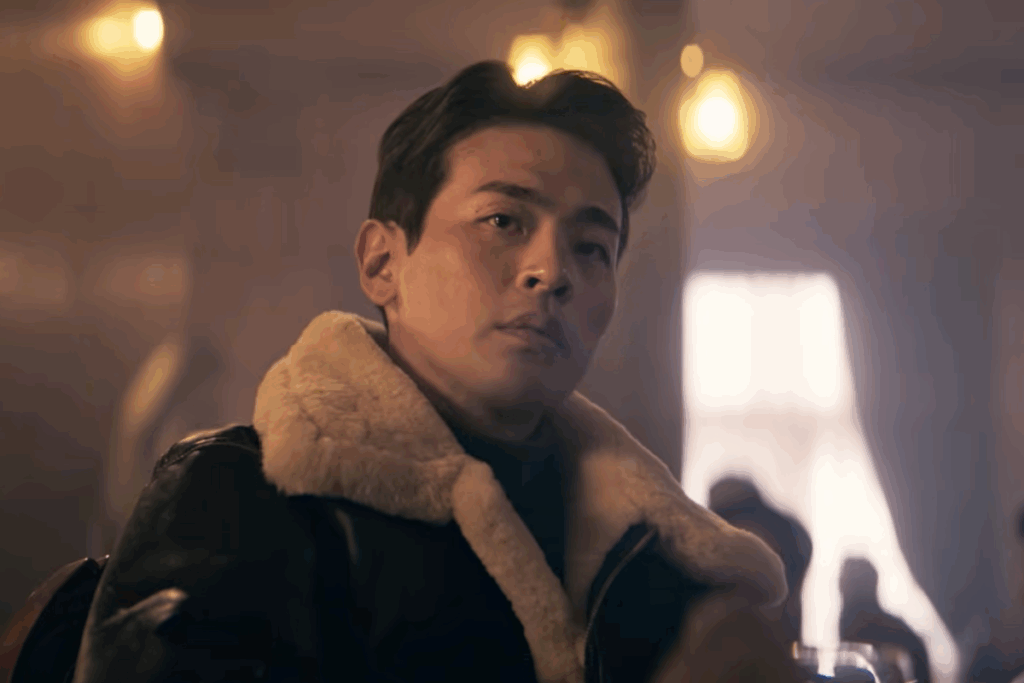 Homem com traços orientais e expressão séria em ambiente interno com iluminação suave e pessoas desfocadas ao fundo