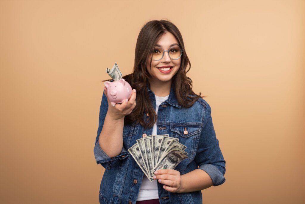 Jovem com cabelo curto, usando óculos redondo preto com notas de dinheiro em uma mão e cofre em forma de porco rosa na outra, usando camiseta branca e camisa jeans em fundo bege