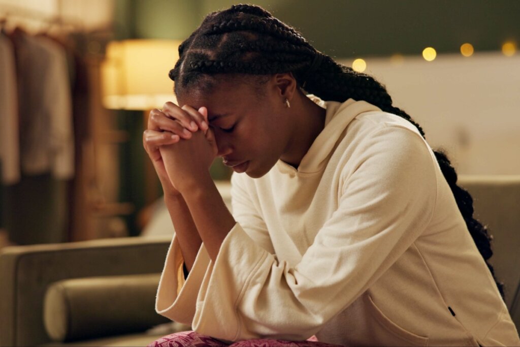 Jovem orando sentada no sofá de olhos fechados e com as mãos cruzadas encostada no rosto