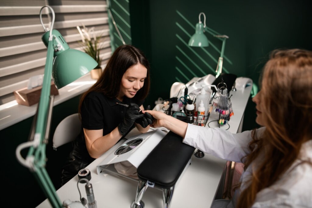 Mulher com cabelo solto preto, usando camiseta preta realizando alongamento de unha em cliente usando camisa branca em estúdio com paredes verdes