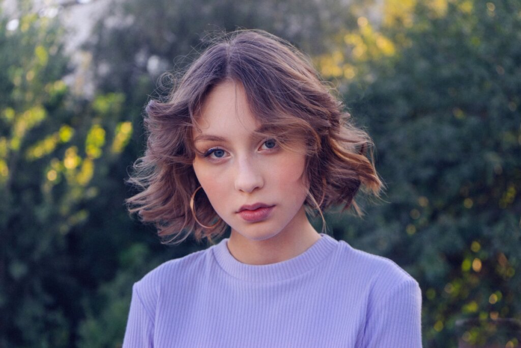 Mulher com cabelo curto com ondas, usando camiseta roxa, posando para foto em um jardim 