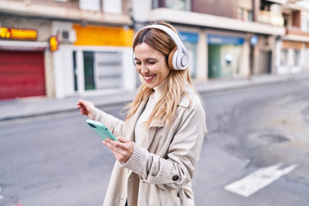 Mulher com cabelo loiro, solto e liso, usando fones de ouvido branco e laranja e camisa e casaco bege com celular na mão andando na rua ouvindo música.