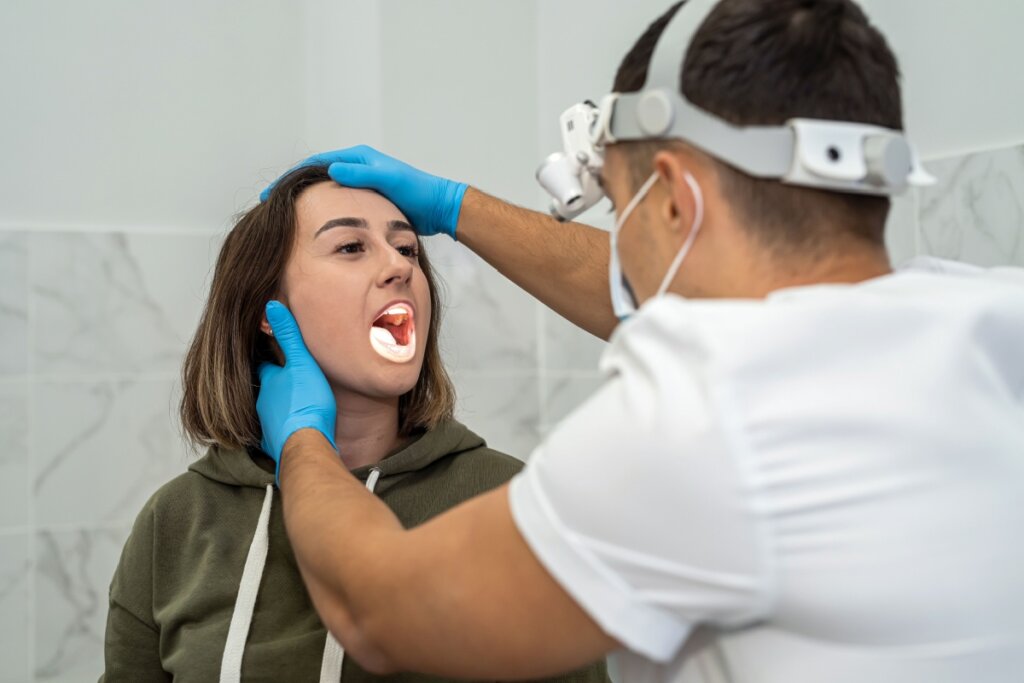 Jovem com o cabelo curto, usando camisa verde, sendo avaliada por médico usando camiseta branca e equipamento iluminando a garganta da paciente