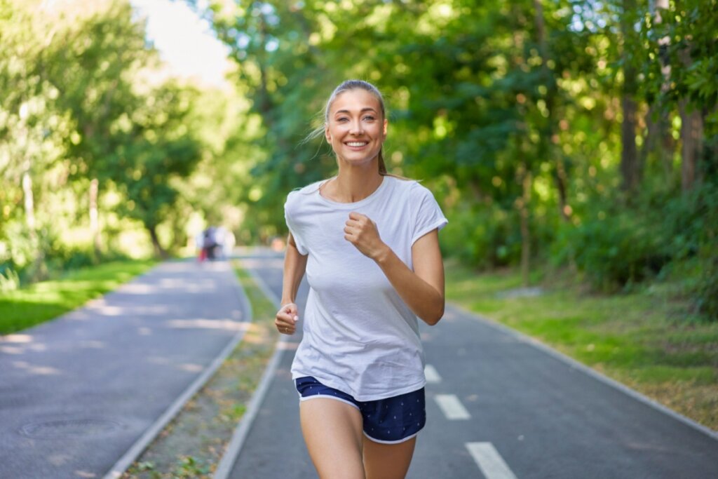 Mulher com o cabelo amarrado, usando camiseta branca e short azul com detalhes brancos correndo no parque