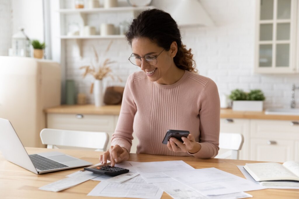 mulher de blusa rosa sentada, sorrindo e segurando o celular. na mesa, há notebook e papéis espalhados 