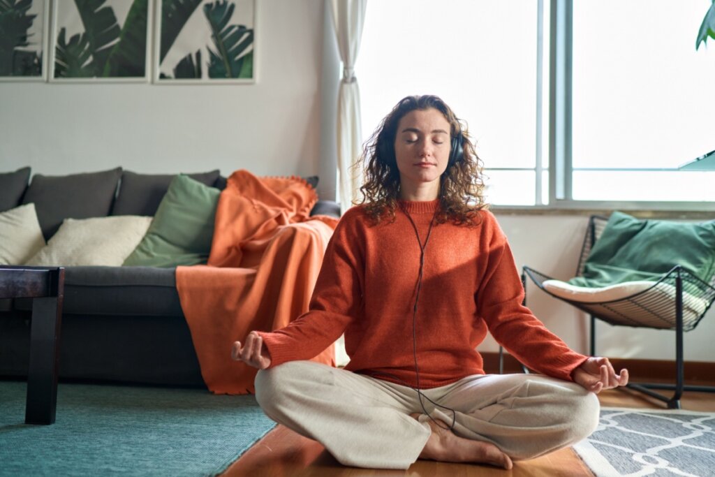 Mulher sentada no chão meditando com fone de ouvido, usando blusa de manga longa laranja e calça bege sentada na sala de estar