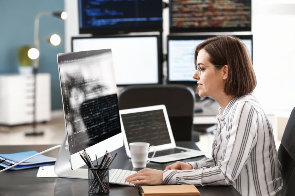 Mulher com cabelo curto, usando camisa de manga longa listrada em tons de cinza trabalhando em dois computadores em escritório