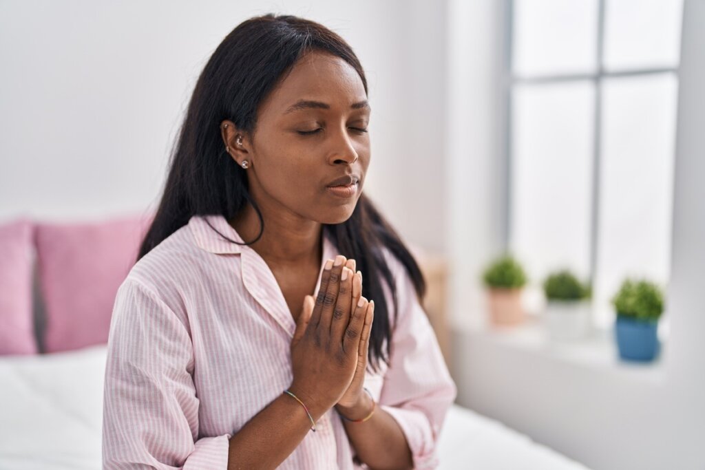 Mulher de olhos fechados e mãos unidas em oração dentro de um quarto iluminado