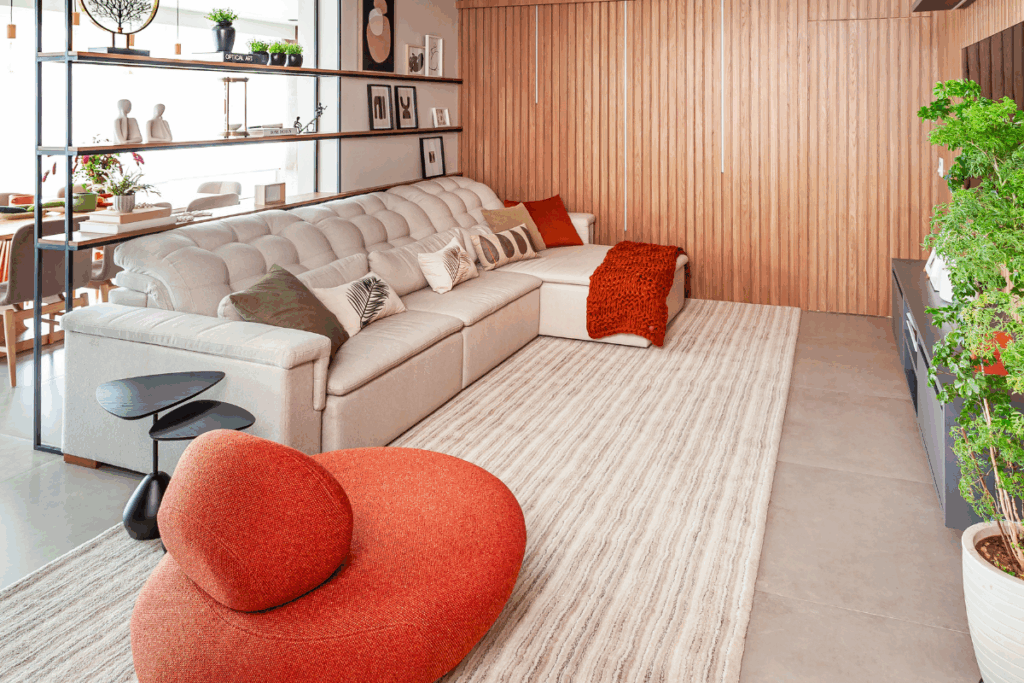 Sala com parede de madeira, sofá bege com almofadas e manta laranja, com poltrona orgânica laranja redonda ao lado do sofá