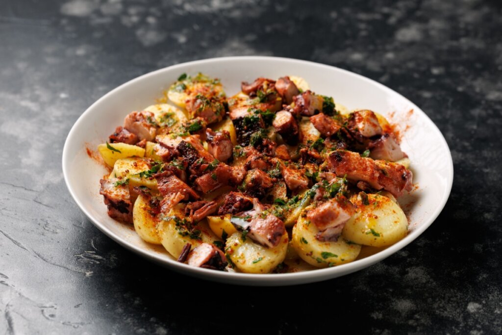 A imagem mostra um prato com pedaços de polvo acompanhados de batatas cozidas e temperos verdes. A preparação aparece em um prato branco, com aparência de prato quente e bem temperado.