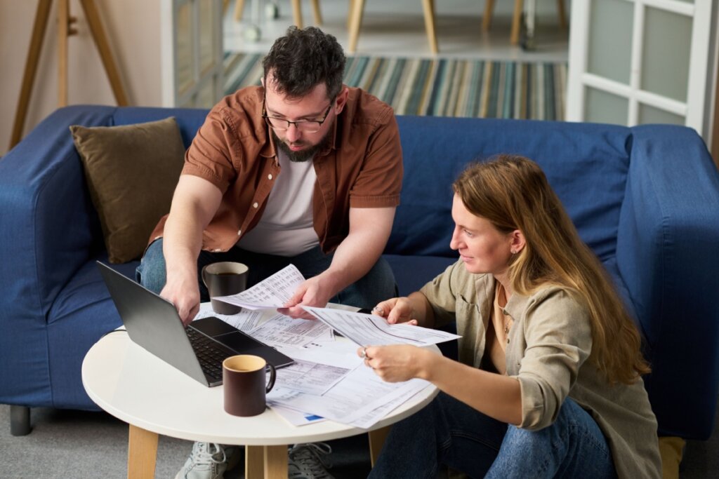 Homem sentado em um sofá e uma mulher sentada no chão, em frente a uma mesa, olhando para notebook e mexendo em papéis