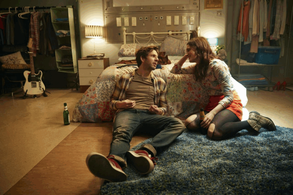 Casal conversando sentado no chão encostado em cama de casal em quarto de hospital, a mulher tem o cabelo longo, solto, está usando uma camiseta estampada, uma saia laranja e meia preta e bota marrom e o homem tem o cabelo curto, está usando uma camiseta listrada e uma camisa xadrez e calça jeans e tênis marrom