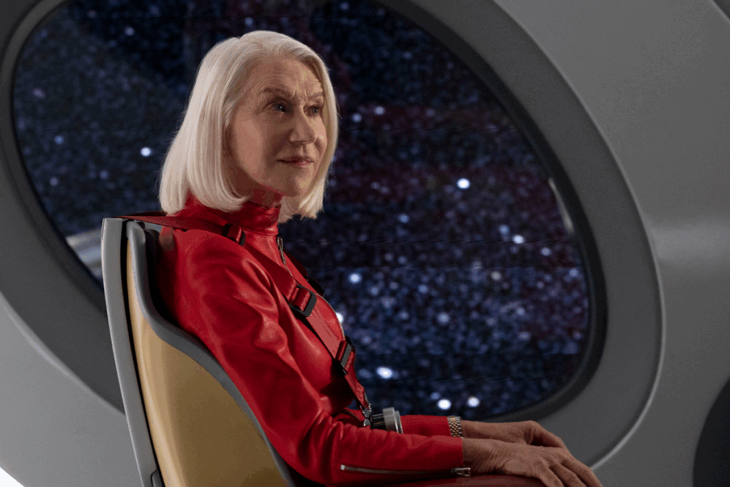 Mulher com cabelo grisalho e curto, usando uniforme vermelho sentada em cadeira em nave, com uma janela ao fundo mostrando o espaço