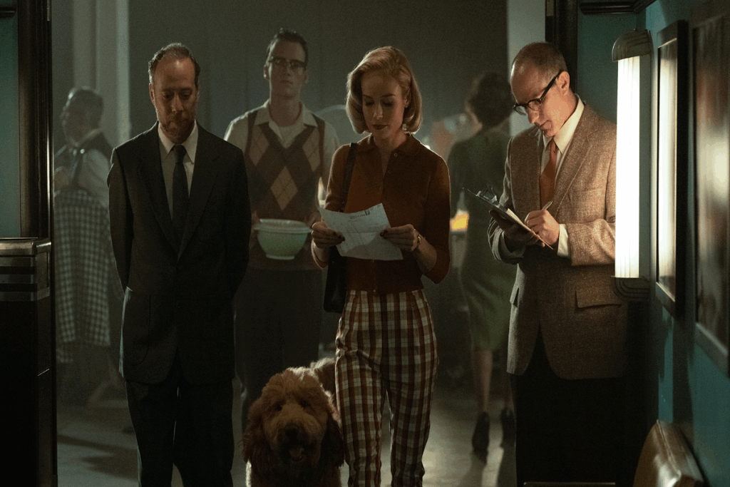 Mulher com cabelo curto, usando camisa marrom e calça xadrez andando lendo papel com homens ao lado vestindo roupas de época e um cachorro com o pelo marrom ao lado da mulher