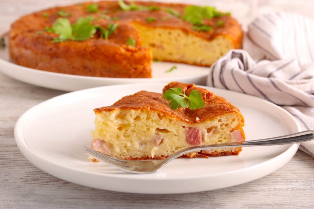 A imagem mostra uma torta salgada assada, com aparência bem fofinha e dourada por fora. Um pedaço já foi cortado e servido no prato, revelando o recheio cremoso com presunto e queijo. A torta está decorada com folhas verdes por cima