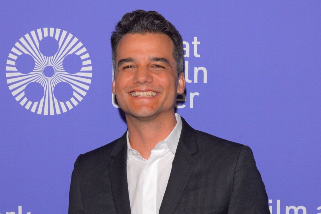 wagner moura sorrindo. ele usa camisa branca e terno preto. fundo da imagem é azul