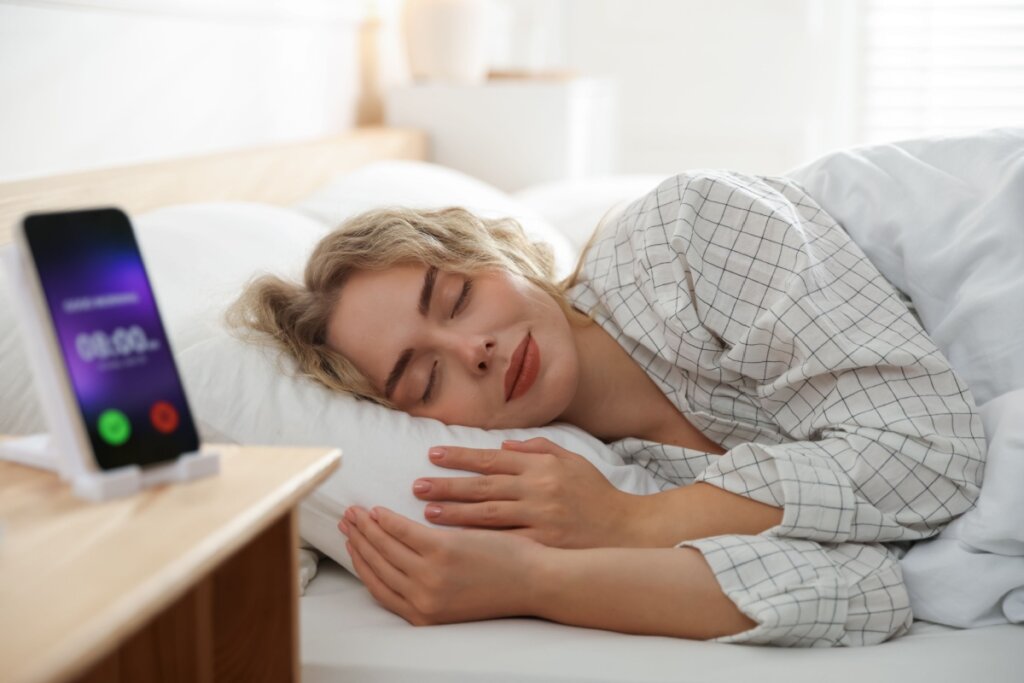 Mulher dormindo tranquilamente na cama ao lado de celular com alarme em ambiente claro