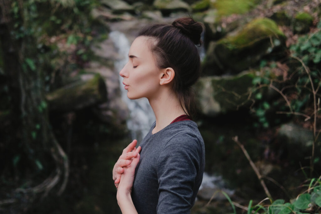 Mulher de olhos fechados e mãos no peito, com o cabelo preso em coque e usando blusa cinza com uma cachoeira ao fundo