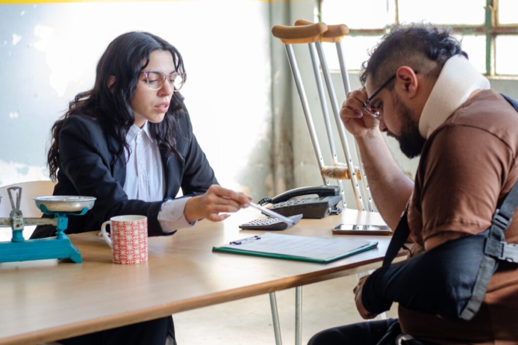 Uma advogada ou especialista em seguros, vestindo blazer preto, está sentada à mesa conversando com um homem que sofreu múltiplos ferimentos. O homem usa um colar cervical, tem o braço esquerdo em uma tipóia e muletas estão apoiadas ao seu lado. A mulher aponta com uma caneta para um documento em uma prancheta sobre a mesa, enquanto o homem, com expressão de preocupação, apoia a mão na testa. Sobre a mesa, veem-se uma balança antiga de metal azul, uma caneca e um telefone fixo.