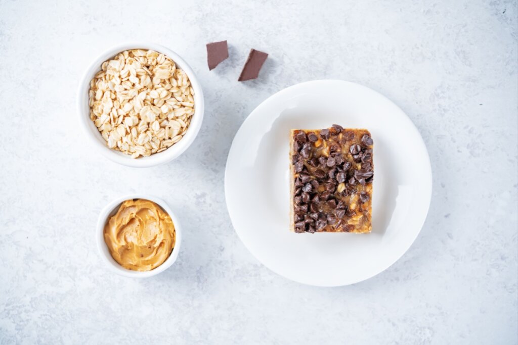Vista superior de uma composição culinária sobre um fundo cinza claro marmorizado. À direita, um prato branco redondo contém uma barra retangular de aveia e pasta de amendoim, generosamente coberta com gotas de chocolate amargo. À esquerda, dois pequenos bowls brancos contêm os ingredientes principais: o de cima está cheio de aveia em flocos e o de baixo contém pasta de amendoim cremosa. Entre o prato e os bowls, há dois pequenos pedaços de chocolate em barra..