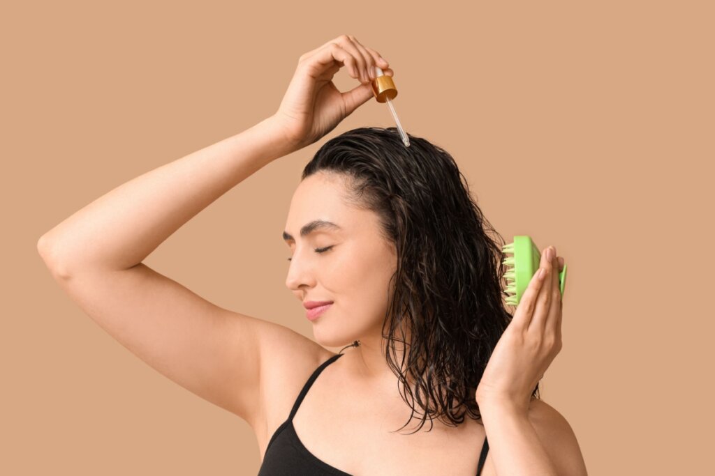 Mulher segura um conta-gotas de vidro acima da cabeça com a mão direita, aplicando um líquido transparente no couro cabeludo. Com a mão esquerda, ela segura uma escova de massagem capilar de silicone verde contra a parte de trás da cabeça. Ela veste uma regata preta de alças finas. A iluminação é suave e uniforme, realçando a textura úmida de seu cabelo