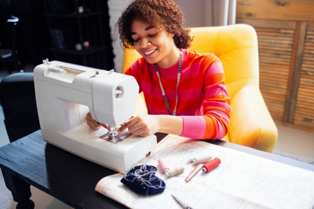 Uma jovem negra de cabelos cacheados sorri enquanto opera uma máquina de costura branca sobre uma mesa de madeira escura. Ela veste uma blusa de listras horizontais em tons de rosa e laranja e possui uma fita métrica pendurada no pescoço. À sua frente, há moldes de papel, carretéis de linha coloridos, um abridor de casas e um almofadinha de agulhas. Ela está sentada em uma poltrona amarela vibrante em um ambiente aconchegante e bem iluminado. A cena transmite criatividade, foco e satisfação com o trabalho manual.