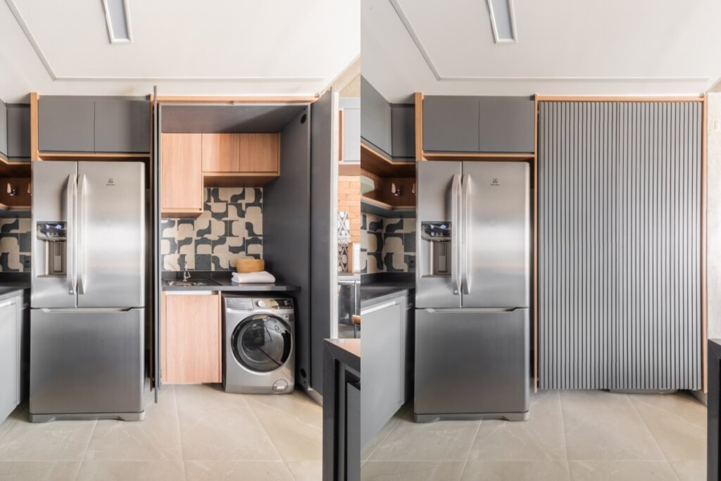 Montagem com duas fotos de uma área de serviço compacta e planejada. À esquerda, o compartimento está aberto, revelando uma máquina de lavar prata, um tanque embutido em bancada de pedra preta, armários de madeira clara e um revestimento de azulejos com formas geométricas em preto e branco. À direita, o mesmo espaço aparece fechado por uma porta de correr cinza com acabamento ripado, que se alinha aos armários superiores e à geladeira de aço inoxidável ao lado, ocultando totalmente a lavanderia.