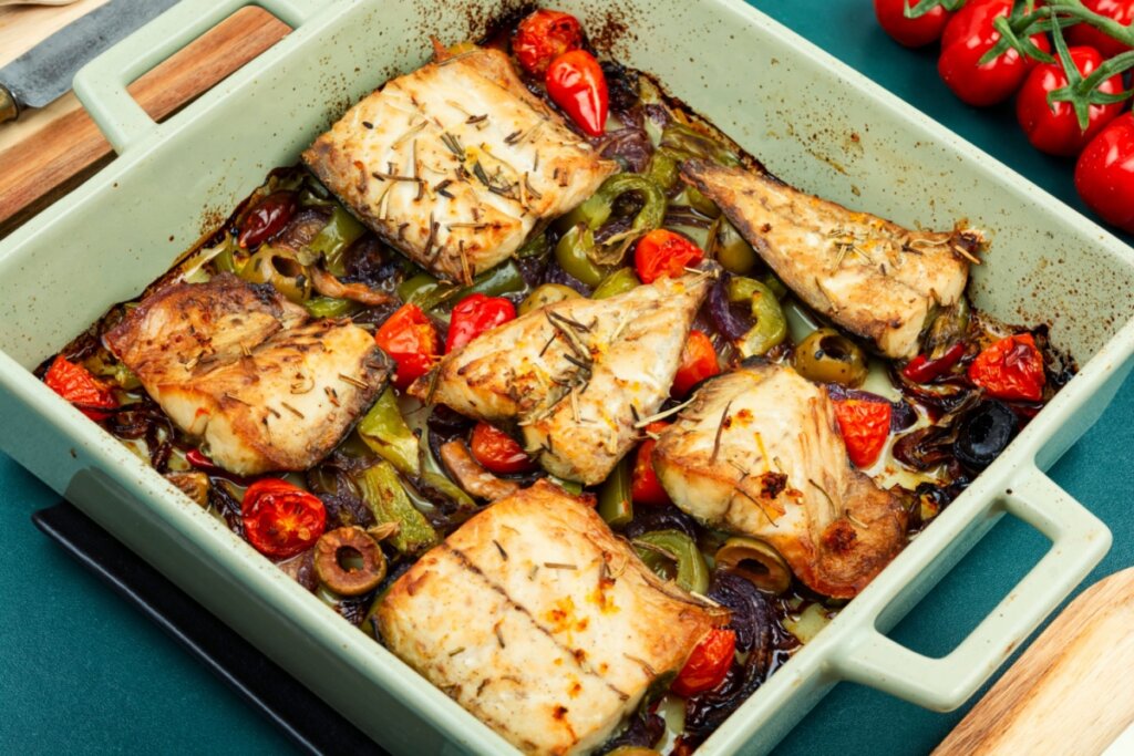 Vista superior de uma assadeira de cerâmica verde clara contendo seis filés de sarda assados. Os peixes estão dispostos sobre uma cama de legumes coloridos que inclui tomates-cereja vermelhos, rodelas de pimentão verde, fatias de cebola roxa e azeitonas verdes. O prato está temperado com ervas secas e páprica. No canto superior direito, fora da assadeira, observa-se um ramo de tomates frescos sobre uma superfície verde.