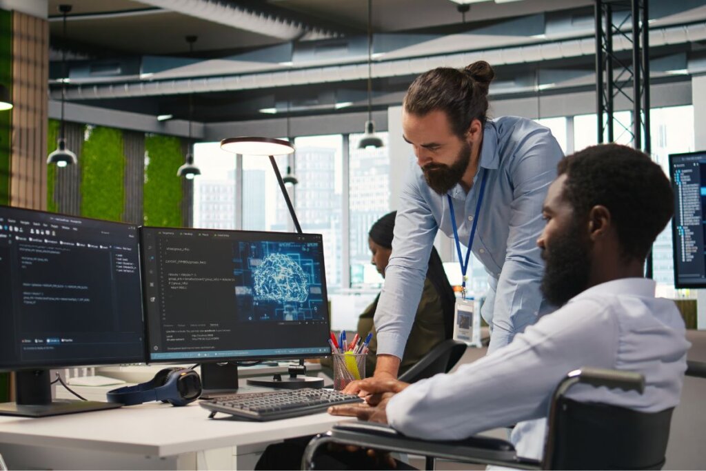 Homem em pé ao lado de outro sentado, ambos olhando para a tela de um computador em um escritório