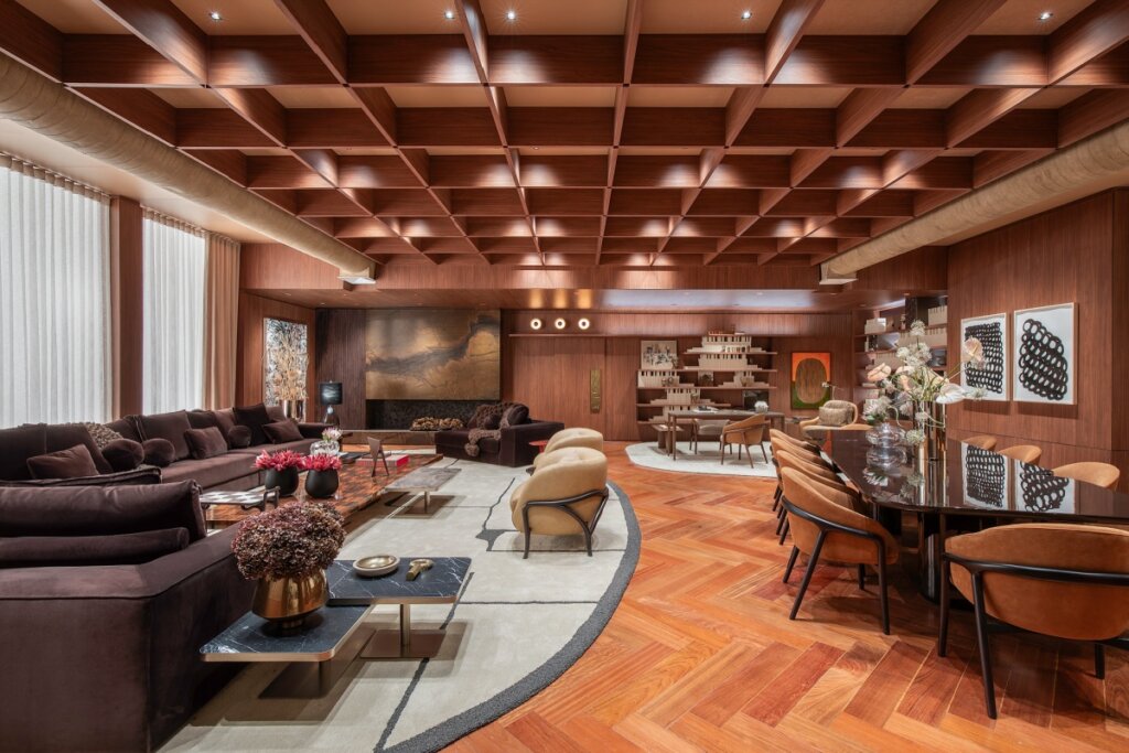 Fotografia de uma sala integrada luxuosa com piso de madeira em padrão espinha de peixe e teto de madeira em grelha profunda. À esquerda, um grande sofá de veludo marrom escuro; à direita, uma mesa de jantar longa e preta com cadeiras estofadas em tom de terracota. O espaço é amplo, conectando a área de estar com um escritório ao fundo, mantendo uma paleta de cores terrosas e materiais naturais.