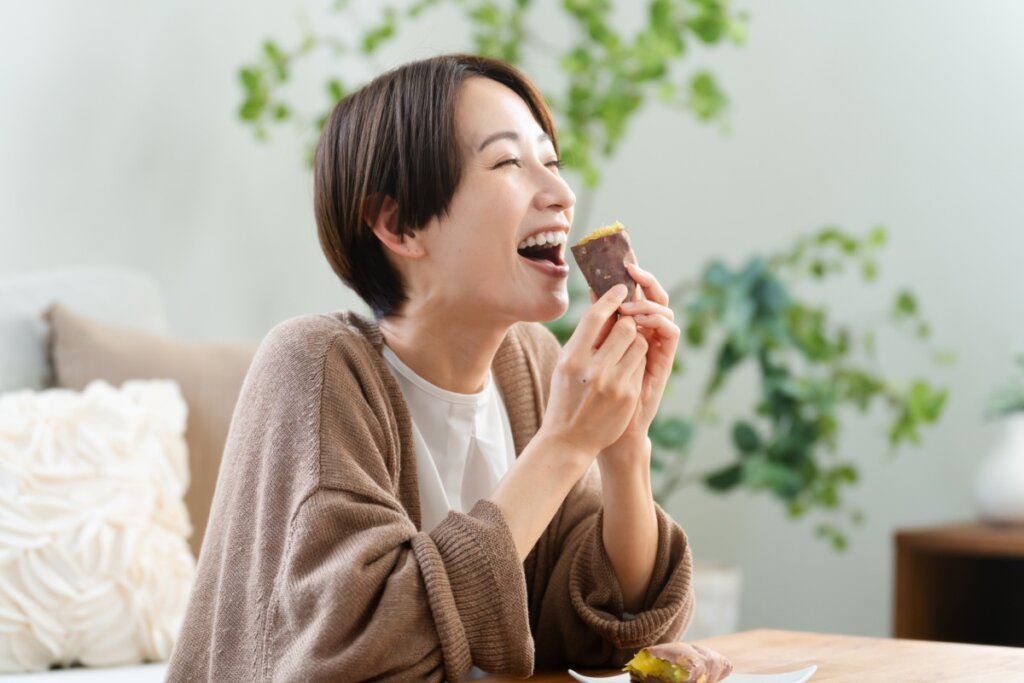 Mulher de cabelos curtos sorrindo alegremente enquanto segura um pedaço de batata-doce cozida. Ela veste um cardigã marrom e está em um ambiente doméstico acolhedor com plantas ao fundo.