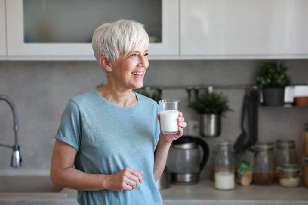 Uma mulher madura sorridente com cabelos curtos e grisalhos está em uma cozinha moderna segurando um copo de leite. Ela veste uma camiseta azul clara e olha para o lado com uma expressão alegre. Ao fundo, vê-se uma bancada de cozinha organizada com utensílios e potes.