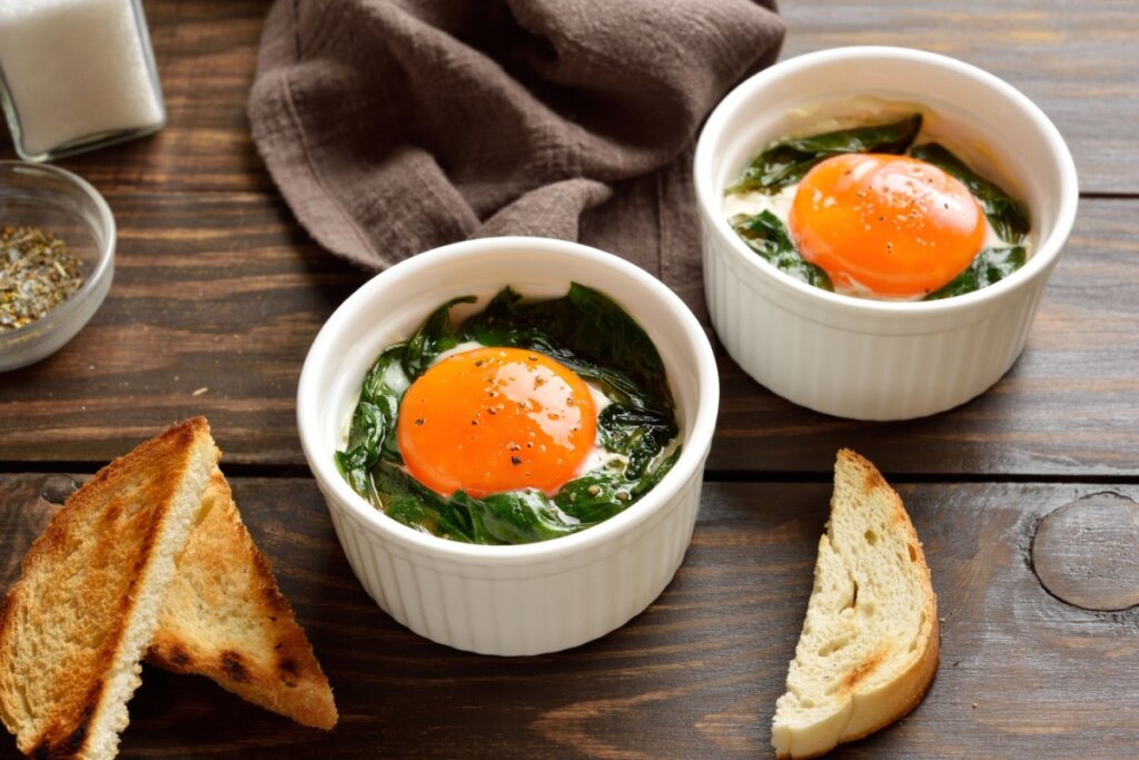 Ramequins com ovos em cocotte sobre espinafre, acompanhados de torradas em mesa de madeira