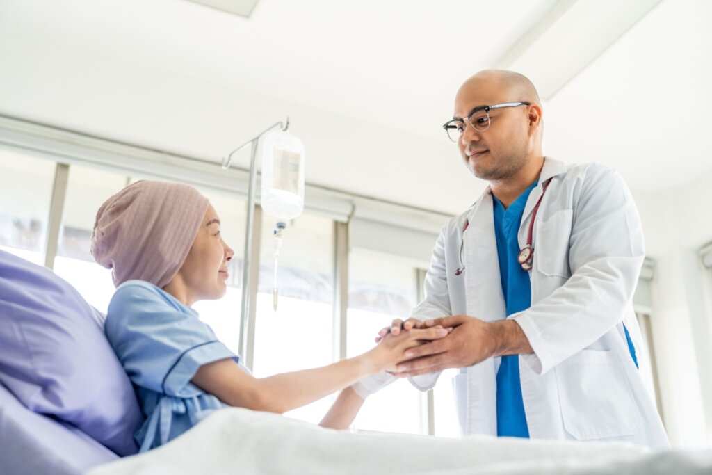 Uma mulher jovem usando um lenço rosa na cabeça está deitada em um leito hospitalar, sorrindo suavemente para um médico. O médico, um homem calvo de óculos e jaleco branco, segura as mãos da paciente em um gesto de apoio e conforto. Ao fundo, vê-se um suporte com soro e janelas amplas que iluminam o quarto de hospital. A cena transmite empatia e cuidado humanizado.