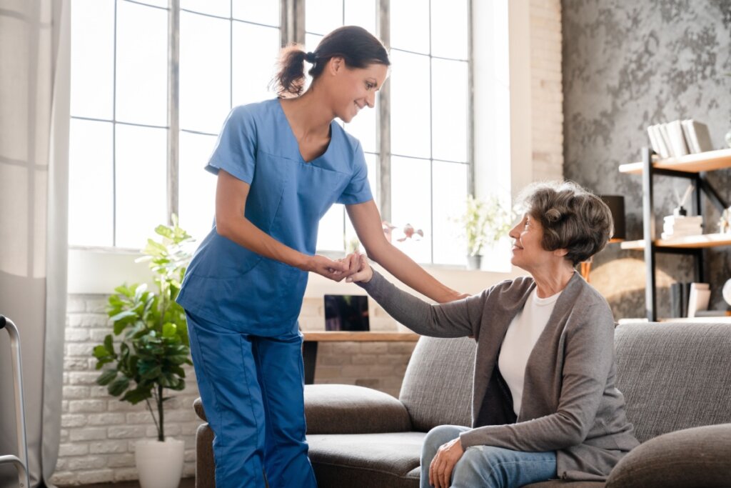 Fotografia de uma enfermeira sorridente, vestindo uniforme azul (scrubs), ajudando uma mulher idosa a se levantar de um sofá cinza. A enfermeira segura delicadamente as mãos da senhora, que retribui o olhar com confiança. O ambiente é uma sala bem iluminada por grandes janelas ao fundo, transmitindo uma sensação de acolhimento e cuidado humanizado.