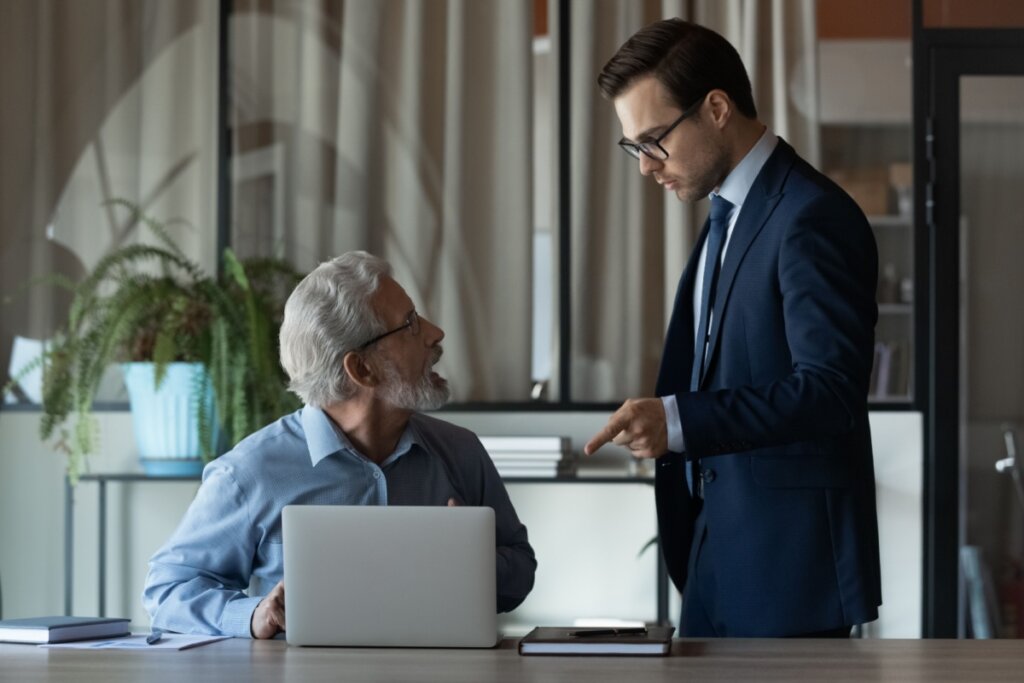 Fotografia de uma interação tensa em um escritório. À direita, um homem jovem de terno azul escuro e óculos está de pé, apontando o dedo de forma assertiva para um notebook sobre a mesa. À esquerda, um homem mais velho de cabelos brancos e camisa social azul está sentado, olhando para cima com uma expressão de surpresa ou defensiva. O ambiente possui tons neutros, com uma planta ao fundo e divisórias de vidro.