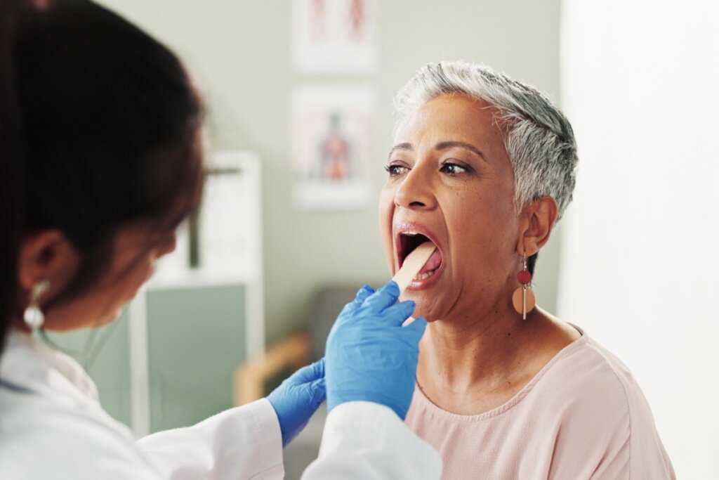 Uma médica, de costas para a câmera e vestindo jaleco branco e luvas azuis, examina a garganta de uma paciente idosa com cabelos grisalhos curtos. A paciente está com a boca aberta e a médica usa um depressor de língua de madeira. A paciente olha para a médica com uma expressão neutra e atenta. O ambiente é um consultório médico com um pôster de anatomia visível ao fundo.