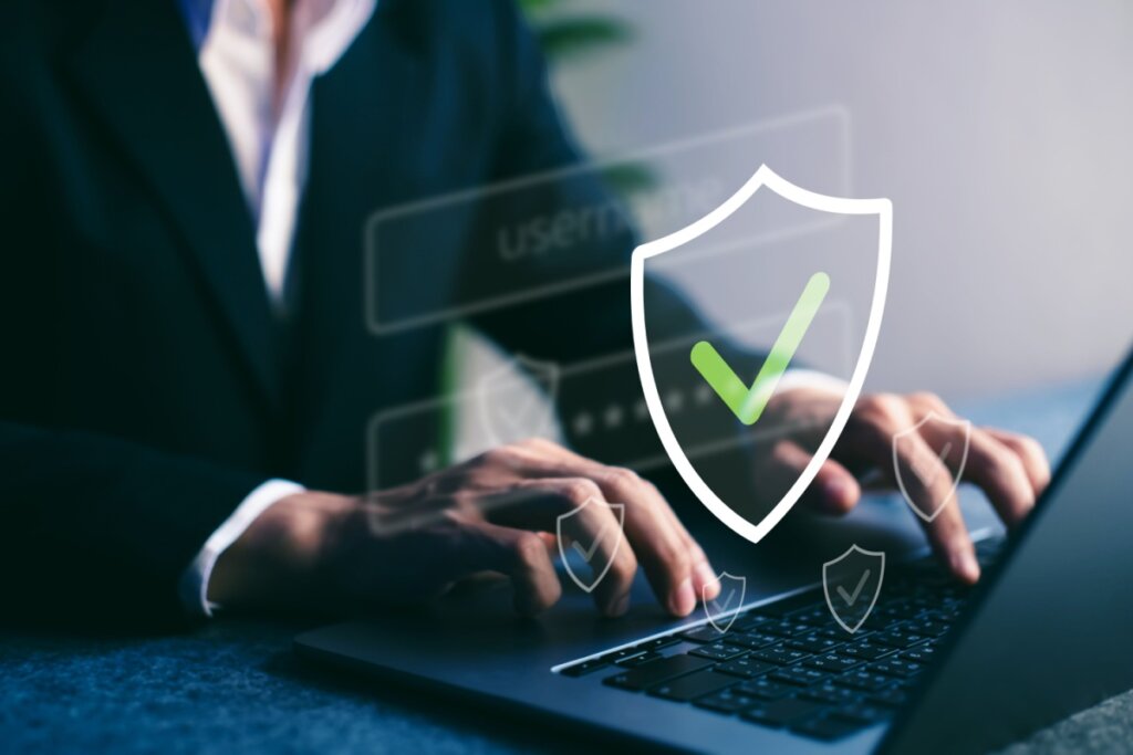 Close-up das mãos de uma pessoa de terno usando um notebook. Sobre a imagem, há ícones digitais flutuantes de escudos, destacando um escudo central branco com um check (verificado) verde, simbolizando proteção de dados e segurança digital