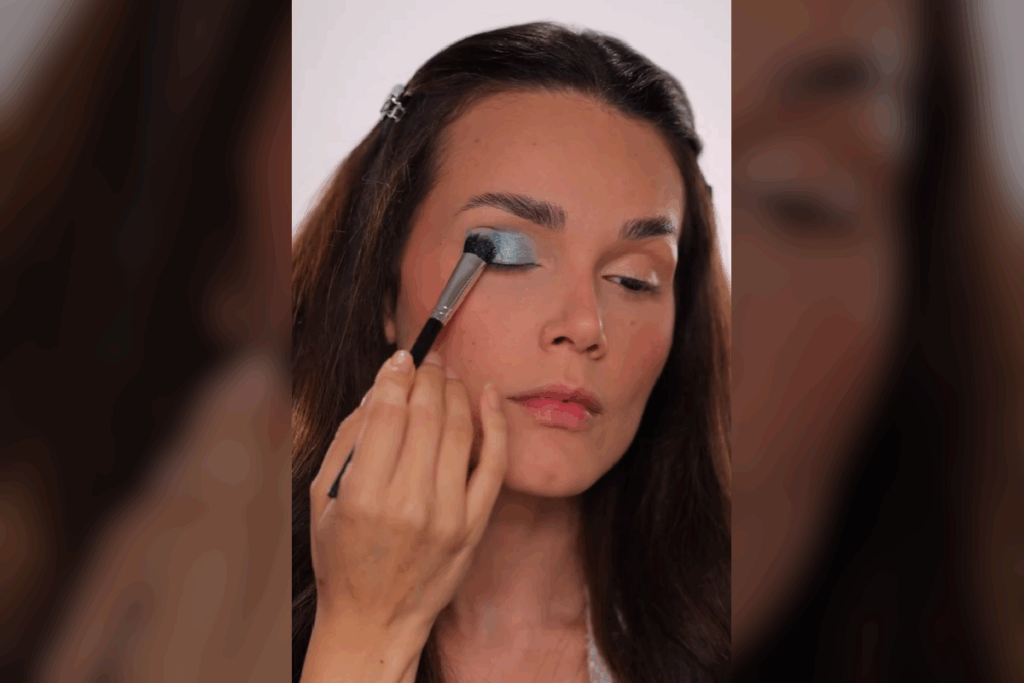 Close do rosto de uma mulher aplicando uma sombra azul-celeste metálica no centro da pálpebra móvel com um pincel de cerdas densas. A iluminação destaca o brilho acetinado da maquiagem, criando um ponto de luz no olhar.