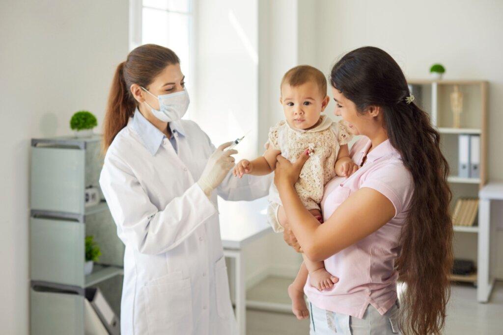 Fotografia em ambiente clínico iluminado, mostrando uma médica pediatra de jaleco branco, máscara e luvas, segurando uma seringa preparada para vacinação. À direita, uma mulher com cabelos longos sorri enquanto segura um bebê no colo, que olha atentamente para a câmera. O cenário ao fundo é composto por prateleiras organizadas e plantas decorativas, transmitindo uma sensação de cuidado e segurança em um consultório médico.