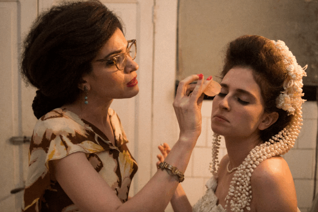 Duas mulheres em cena de época, uma delas vestida de branco com coroa de flores, sendo maquiada pela outra