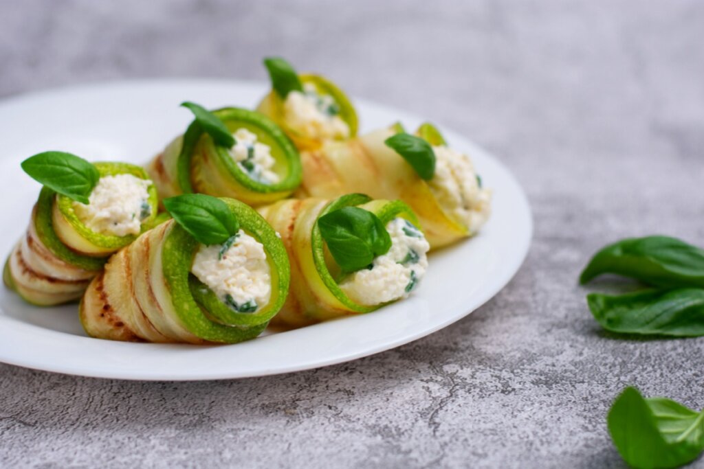 A imagem mostra um prato com rolinhos de abobrinha grelhada recheados com um creme claro. As fatias de abobrinha estão enroladas de forma delicada, criando uma apresentação elegante. Cada porção é finalizada com folhas de manjericão