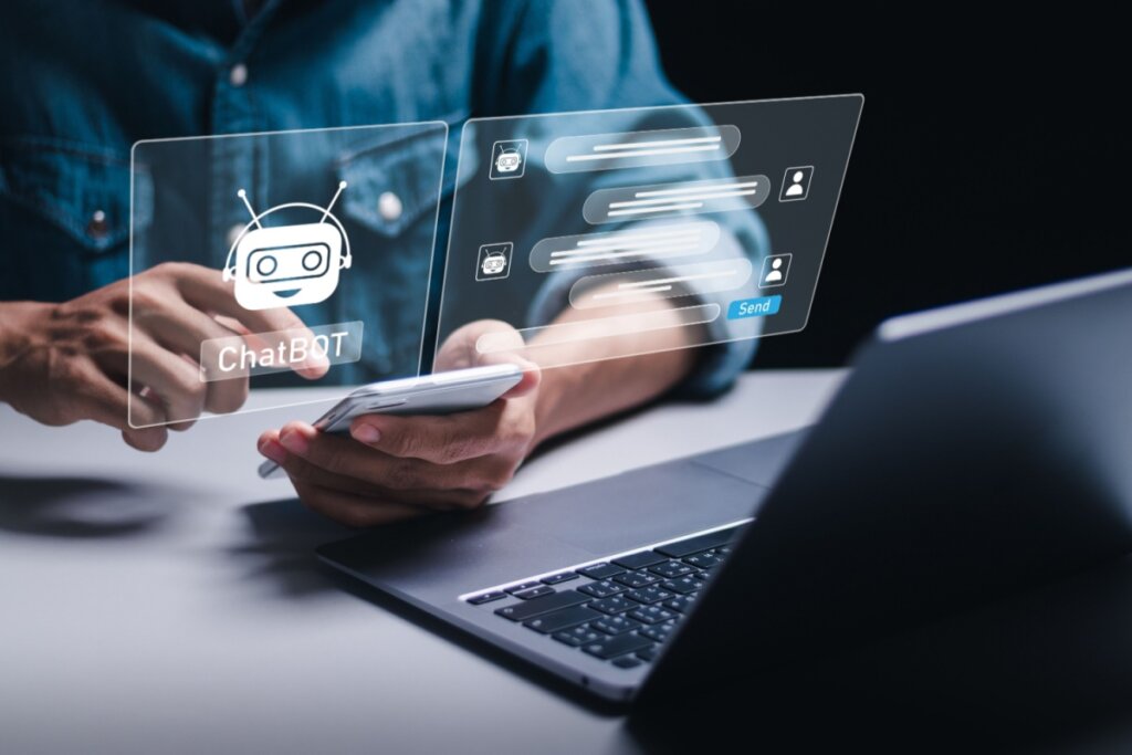 Mãos mexendo em um celular, em frente a um notebook, com a ilustração de uma conversa com chatBOT acima 