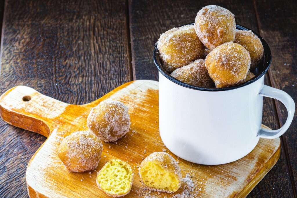 Bolinho de chuva com laranja servido em caneca branca com fundo preto, em cima de tábua de madeira com bolinhos ao redor e um cortado ao meio em cima de mesa de madeira 