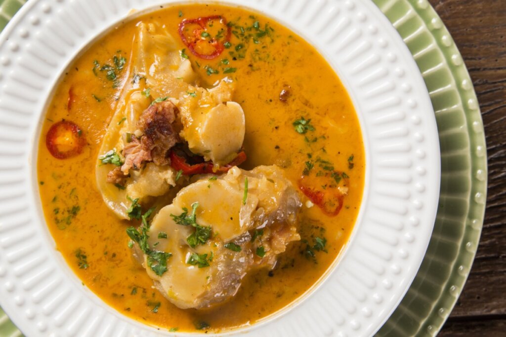 Uma fotografia de um prato fundo branco com bordas trabalhadas em relevo, servido sobre um prato raso verde-claro. Dentro do prato, há um caldo de mocotó denso e alaranjado, contendo pedaços grandes de cartilagem cozida e carnes suculentas. O prato é finalizado com salsinha picada e pequenos pedaços de pimenta vermelha.